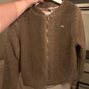 Beige teddyjacka från Aim'n - Mysig teddyjacka från Aim'n i beige färg med fluffig yta och rund hals. Jackan har dragkedja framtill och är tillverkad i 100% polyester. Perfekt för dig som gillar chill och comfy stil. Långärmad och enkel att matcha med jeans eller leggings.