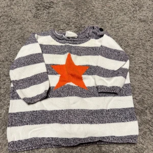 Randig stickad tröja med stjärna Kappahl storlek 62 - Superfin stickad tröja från Kappahl i storlek 62. Tröjan är vit och grå med breda ränder och har en orange stjärna framtill. Långärmad modell med knappar på axeln för enkel påklädning. Perfekt för kyligare dagar.