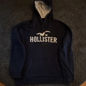 Mörkblå Hollister hoodie med logga - Snygg mörkblå hoodie från Hollister med stor vit broderad logga och text på bröstet. Tröjan har huva med ljus insida, snörning och en klassisk känguruficka framtill. Perfekt för dig som gillar en avslappnad och nonchalant stil