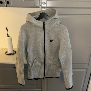 Grå hoodie från Nike med dragkedja - Snygg grå hoodie från Nike i storlek XS. Tröjan har hel dragkedja framtill, två fickor och svart Nike-logga på bröstet. Mjuk och skön med huva och svarta detaljer längs dragkedjan och ärmsluten. Perfekt för chill eller träning. (passar s)