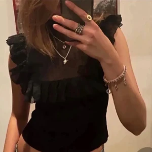 Svart blus med volangdetaljer - Säljer en svart blus med transparent mesh upptill och snygga volangdetaljer längs axlar och bröst. Blusen är ärmlös och har en croppad passform som ger en trendig look. Perfekt för dig som gillar att sticka ut med coola detaljer och stilren design.