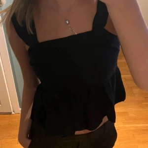 Svart volangtop från Kappahl - Säljer en svart linnetopp från Kappahl i storlek S. Toppen har breda axelband, smockad överdel och volang nedtill som ger en söt och trendig look. 