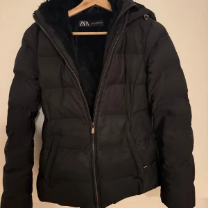 Fin svart pufferjacka från Zara - En väldigt fin, svart pufferjacka från ZARA – både varm och stilren. Den är lätt, har mjukt foder på insidan och en huva som skyddar mot kylan. Klassisk passform med två fickor på sidorna och stilren look. Perfekt för höst och vinter när du vill hålla dig varm utan att tumma på stilen. En jacka som passar till allt, bekväm och alltid snygg!