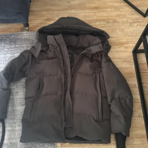 Svart pufferjacka från Canada Goose - Säljer en svart pufferjacka från Canada Goose med huva och diskret logga på ärmen. Jackan har dragkedja och knappar framtill, två stora fickor och ribbade muddar vid ärmsluten. Perfekt för kalla vinterdagar och riktigt snygg streetstil. Pris kan diskuteras