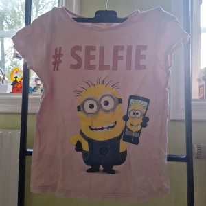 Rosa Minions t-shirt  - Söt rosa t-shirt från Minions med tryck av en minion som tar selfie och texten #SELFIE i glittrigt rosa. T-shirten har korta ärmar och är gjord i mjuk bomull, perfekt för dig som älskar roliga prints och filmkaraktärer.