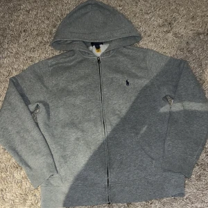 Grå hoodie från Polo Ralph Lauren - Klassisk grå hoodie från Polo Ralph Lauren med dragkedja framtill, huva och två fickor. Tröjan har broderad logga på bröstet och är tillverkad i mjuk bomullsmix. Perfekt för chill dagar eller när du vill ha en enkel och stilren look.