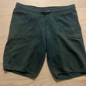 Mörkgröna shorts Peak Performance - Mörkgröna shorts från Peak Performance med snyggt tryck längs benet. De har en bekväm passform, elastisk midja med snörning och två stora fickor framtill. Perfekta för chill dagar eller träning. Materialet känns mjukt och sportigt.