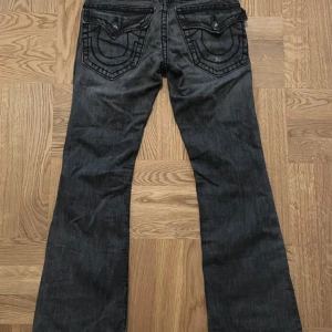 True Religion svarta bootcut jeans - Säljer ett par svarta jeans från True Religion, modell Billy Super T. Jeansen har markerade sömmar, stora bakfickor med lock och knappar samt bootcut passform. Tillverkade i USA och i klassisk denim med cool tvättad look.           Midja- 40cm Ytterben-100cm benöppning-23cm