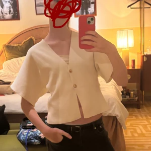 Fin blus - Säljer en beige blus med v-ringning och två snygga guldfärgade knappar framtill. Blusen har korta, vida ärmar och är croppad i modellen. Materialet känns mjukt och har lite struktur, perfekt för dig som gillar minimalistisk och clean stil.