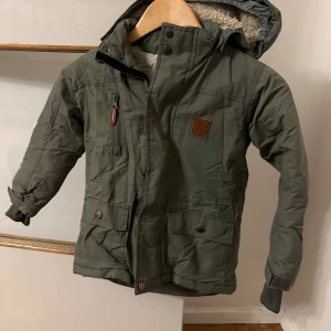 Grön parkajacka från Kuling - Säljer en grön parkajacka från Kuling med mysigt teddyfoder på insidan och huva. Jackan har flera fickor med knappar, dragkedja och muddar vid ärmslut. Perfekt för kalla dagar och har snygga detaljer som brunt märke med K på bröstet.