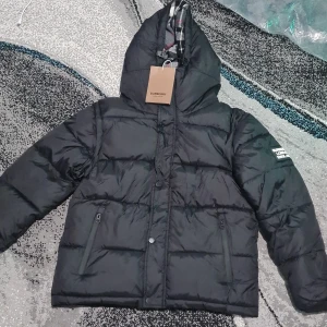 Svart Burberry pufferjacka med avtagbara ärmar - Svart pufferjacka från Burberry med huva och klassiskt rutigt foder. Jackan har avtagbara ärmar så den kan användas som väst. Två fickor med dragkedja framtill och Burberry-logga på ärmen. Perfekt för dig som gillar att variera din stil.