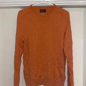 Orange merinoulltröja från Morris - Säljer en snygg orange stickad tröja från Morris i 100% merinoull. Tröjan har rund halsringning, ribbade muddar och en liten broderad logga på bröstet. Perfekt för lager-på-lager under hösten och vintern.