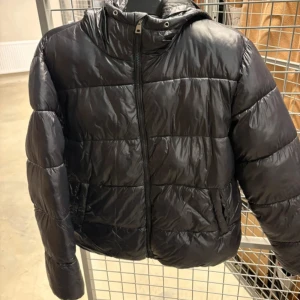 Svart pufferjacka med huva - Svart pufferjacka med huva och dragkedja framtill. Jackan har ett quiltat mönster och är tillverkad i glansig syntet. Perfekt för kalla dagar och har en clean, modern look. Långärmad och med praktiska fickor på sidorna.