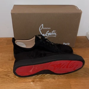 Christian Louboutin skor - Svarta sneakers från Christian Louboutin med ikonisk röd sula och snygg signatur på undersidan. Skorna har rund tå, platt sula och är tillverkade i mjuk mocka med svarta snören. Perfekt för dig som vill ha en lyxig och stilren look. Storlek 44 passar även 43, kan liven gå ner i pris vid snabb affär
