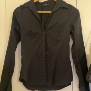 Svart figursydd skjorta med krage - Svart skjorta med klassisk krage och lång ärm. Figursydd modell med knäppning framtill. Perfekt för en clean och stilren look. Tunn och lätt i materialet, passar bra till både jeans och kostymbyxor.