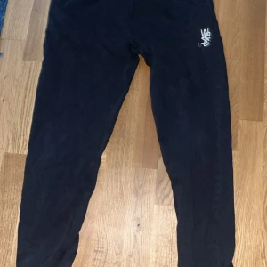 Svarta mjukisbyxor med broderad logga - Svarta mjukisbyxor med en broderad vit logga på ena höften. Byxorna har en avslappnad passform och en bakficka. Perfekta för chill dagar eller när du vill vara bekväm men ändå se snygg ut.