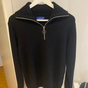 Svart stickad half zip-tröja Jack & Jones - Svart stickad tröja från Jack & Jones med ribbad hög krage och snygg half zip-dragkedja med ringdetalj. Långärmad och i klassisk modell, perfekt för lager-på-lager under kyliga dagar. Tveka inte att höra av dig😁