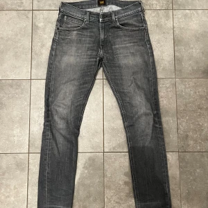 Grå jeans från Lee, slim fit - Snygga grå jeans från Lee med klassisk femficksdesign och diskreta slitningar. Modellen har smal passform och raka ben, perfekt för dig som gillar en stilren look. Jeansen har dragkedja och knappstängning samt Lee-logga på bakfickan. 29/32 i storlek 