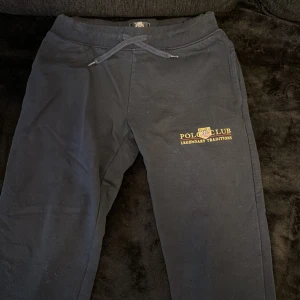 Svarta mjukisbyxor från Vinson Polo Club - Svarta mjukisbyxor från Vinson Polo Club med broderad logga och text i gult på vänster ben. Byxorna har ribbad midja med snörning och är gjorda i mjukt bomullsmaterial. Perfekta för chill eller sportiga dagar.