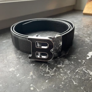 Svart Burberry bälte med B-spänne - Snyggt svart bälte från Burberry med ett stort, silvrigt B-format spänne. Bältet har ett diskret rutigt mönster och är gjort i ett tåligt skinnmaterial. Perfekt accessoar för att lyfta din outfit med en lyxig touch.