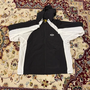 Svart och vit vindjacka från Corteiz - Säljer en svart och vit vindjacka från Corteiz med huva och dragkedja framtill. Jackan har coola vita paneler längs ärmarna och logga på bröstet samt ryggen. Materialet är lätt polyester och jackan har meshfoder på insidan. Perfekt för dig som gillar streetwear.