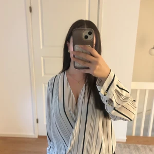 Vit randig omlottblus från Zanzi - Snygg vit omlottblus från Zanzi med svarta ränder. Blusen har lång ärm med guldfärgade knappar vid manschetten och ett v-ringad omlottliv som ger en chill och trendig look. Materialet är mjukt och känns som polyester.