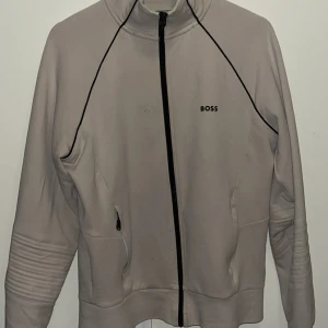 Beige zipuptröja från BOSS - Snygg beige zipuptröja från BOSS med svart dragkedja och diskret logga på bröstet. Jackan har svarta detaljer längs axlarna, ribbade partier vid ärmarna och praktiska fickor med dragkedja. Perfekt för dig som gillar stilren och sportig look.