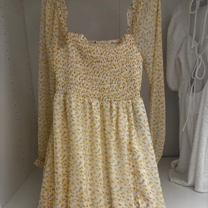 Blommig långklänning med smock - Supersöt långklänning i vitt med små gula blommor, smockad överdel och långa, luftiga ärmar. Klänningen har en slits framtill och är gjord i ett lätt, flowy material som känns perfekt för varmare dagar. Endast använd en gång!