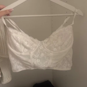Vit spetskorsett från H&M Divided - Säljer en vit korsett-topp från H&M Divided i storlek M. Toppen har tunna axelband, spetsdetaljer och dekorativa sömmar framtill. Baksidan har hakknäppning för extra support. Aldrig använd 💕😇