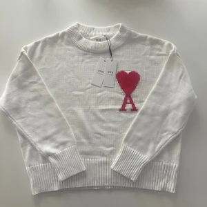 Vit stickad tröja Ami Paris S - Vit stickad tröja från Ami med ribbad rund hals och muddar. På bröstet finns ett stort rött hjärta och bokstaven A i ett coolt mönster. Riktigt stilig nu till vinter o höst!