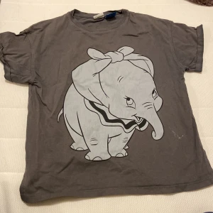 Grå Disney Dumbo t-shirt från Zara - Säljer en grå t-shirt från Zara med stor print av Dumbo på framsidan och texten 'look at me fly.' på baksidan. T-shirten har korta ärmar och är gjord i mjuk bomull. Perfekt för dig som älskar Disney och vill ha en chill och unik look.