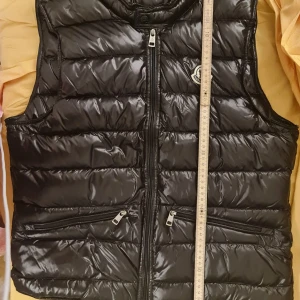 Svart dunväst från Moncler, S - Svart, glansig dunväst från Moncler med quiltad design och klassisk logotyp på bröstet. Västen har två dragkedjeförsedda fickor framtill och en rak, normal passform. Perfekt för lager-på-lager och har en stilren look med hög krage och hel dragkedja.