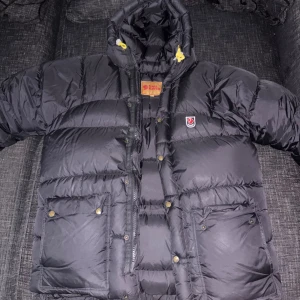 Fjällräven vinterjacka - Storlek S men sitter som XL. Säljer en svart, riktigt fet dunjacka från Fjällräven med huva och gula snören. Jackan har två stora fickor med knappar, dragkedja och klassisk Fjällräven-logga på bröstet. Perfekt när du vill hålla dig varm och ändå se chill ut. PRIS KAN DISKUTERAS‼️