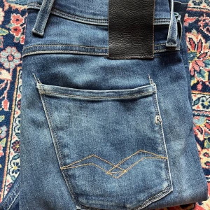 Replay Anbass jeans blå denim - Säljer ett par snygga Replay Anbass jeans i blå denim med klassisk femficksdesign och ljusare tvättade partier. Jeansen har raka ben och detaljerade sömmar på bakfickorna. Perfekt passform och stilren look för dig som gillar kvalitetsjeans. Uppsydda i bena till 30