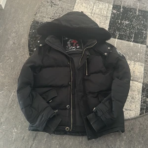 Svart Moose Knuckles dunjacka med luva - Säljer en svart dunjacka från Moose Knuckles med fluffig huva och snygga detaljer som dragkedjor och knappar. Jackan har ribbade muddar, flera fickor och logotyp på ärmen. Perfekt för kalla dagar och har en riktigt schysst streetstil. Riktigt skön vinterjacka