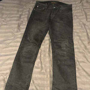 Svarta jeans från Nudie Jeans Co - Svarta jeans från Nudie Jeans Co med klassisk femficksdesign och diskret logga på bakfickan. Jeansen har raka ben och normal passform, tillverkade i slitstarkt jeanstyg. Perfekta för dig som gillar en stilren och enkel look.