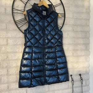 Mörkblå lång dunväst från Attrait - Lång, quiltad dunväst från Attrait Paris i glansig mörkblå färg. Västen har hög krage, dragkedja framtill och ett snyggt rutmönster upptill. Perfekt för lager-på-lager och ger en cool siluett.