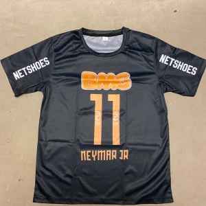 Svart Neymar Jr fotbollströja M - Cool svart fotbollströja med orange tryck, nummer 11 och Neymar Jr på framsidan. Tröjan har korta ärmar, rund hals och är tillverkad i ett lätt, ventilerande polyestermaterial. Sponsorer som NETSHOES och BMG syns tydligt på ärmar och bröst.