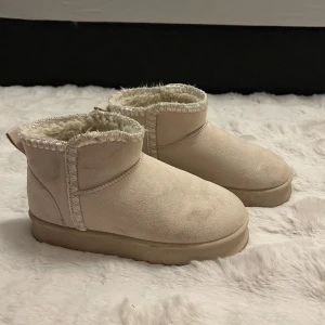 Beiga boots med foder och mocka - Mysiga beiga boots i mocka med fluffigt foder och dekorativa sömmar. Skorna har platt sula och rund tå, perfekta för kalla dagar. Lätta att dra på tack vare den låga modellen och hällan bak.