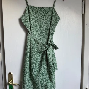 Grön blommig omlottklänning - Söt grön klänning med små vita blommor, smala axelband och omlottknytning i midjan. Lätt och flowy modell som ger en avslappnad vibe, perfekt för varma dagar. Klänningen har en feminin siluett och är knälång.