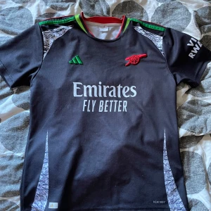 Arsenal svart bortatröja Adidas S - Svart Arsenal bortatröja från Adidas med gröna och röda detaljer vid kragen. Broderat klubbmärke och Adidas-logga på bröstet, samt Emirates Fly Better-tryck. Kortärmad modell i lätt funktionsmaterial med vita mönsterdetaljer på sidorna.