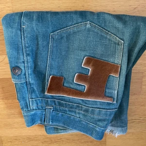 Unika blå jeans med bruna pälsdetaljer - Säljer ett par blå jeans med breda ben och råa kanter nertill. På bakfickorna finns stora bruna pälsapplikationer i form av bokstaven E. Jeansen har klassisk femficksdesign och är tillverkade i denim. Perfekt för dig som vill sticka ut med en cool och annorlunda look. saknar tyvärr knapp!