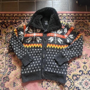 Sjukt unik stickad cardigan - Mysig stickad Cardigan från Rewiew i mörkgrått med vita, orange och gula detaljer. Koftan har stora snöflingemönster, knappar framtill och ett fluffigt svart foder i kragen. Perfekt för kalla dagar och har långa ärmar och ribbade muddar.