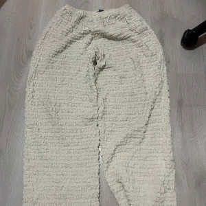 Beiga strukturerade byxor från Zara - Säljer ett par beiga byxor från Zara i storlek S. Byxorna har en unik, strukturerad och rynkad yta med resår i midjan för extra komfort. De är vida i modellen och tillverkade i ett mjukt material som känns skönt mot huden.