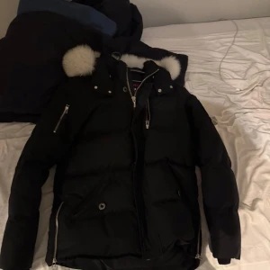 Svart dunjacka Moose Knuckles med päls - Svart dunjacka från Moose Knuckles med fluffig vit fuskpäls på huvan, silverfärgade dragkedjor och logga på ärmen. Jackan har flera fickor med dragkedja och tryckknappar, samt quiltad design för extra värme. Perfekt för kalla vinterdagar. Obs äkta pris går att diskutera då jag inte använder den längre perfek för dig i vintern 