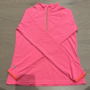 Rosa träningströja från C/FIT - Sportig rosa långärmad träningströja från C/FIT med orange detaljer och half zip framtill. Perfekt för löpning eller gym, tillverkad i mjukt syntetmaterial som andas. Snygg och bekväm passform med reflekterande detaljer på ärmen.