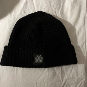 Svart ribbstickad mössa Stone Island - Svart ribbstickad mössa från Stone Island med uppvikt kant och klassisk logopatch framtill. Tillverkad i mjukt material som håller dig varm och ger en clean streetwear-look. Perfekt accessoar för kyliga dagar. Väldigt bra skick, sparsamt använd. 