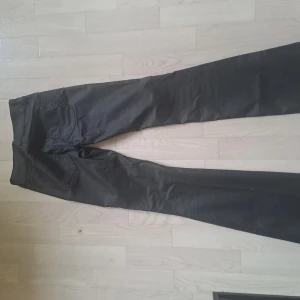 Svarta bootcut-byxor från Nelly - Säljer ett par svarta bootcut-byxor från Nelly med hög midja och snygga fickdetaljer framtill. Byxorna har en lätt glansig finish och är tillverkade i ett syntetmaterial som ger en cool look. Perfekta för dig som gillar trendiga och bekväma byxor.