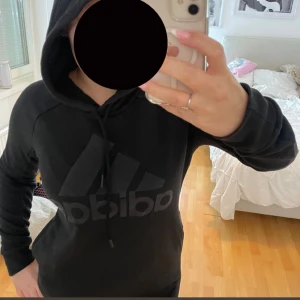Svart hoodie från Adidas med logga - Svart hoodie från Adidas med stor logga framtill och snörning i huvan. Tröjan har lång ärm och klassisk passform, perfekt för chill eller träning. Mjuk och skön i materialblandning med polyester och bomull.