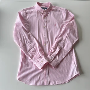 Polo Ralph Lauren Rosa randig skjorta  - Snygg rosa skjorta från Polo Ralph Lauren i slim fit med vita ränder och klassisk button-down krage. Skjortan har broderad logga på bröstet, långa ärmar och knappar framtill. Tillverkad i mjuk bomull för en fräsch och stilren look.